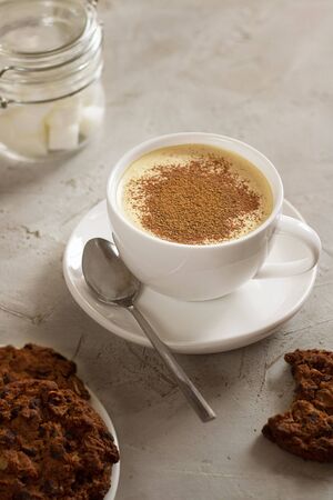 Cappuccino coffee in white cup with tea spoon and american cookies with chocolate drops and defocused sugar bowl on grey table. Cement texture. Breakfast. Cafe. Vertical composition with copy spaceの写真素材