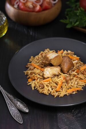 Traditional pilaf with turkey on black plate on dark wooden table with defocused salad, tomatoes, greens and oil. Eastern, uzbek dish made from meat and rise. Vertical.の写真素材