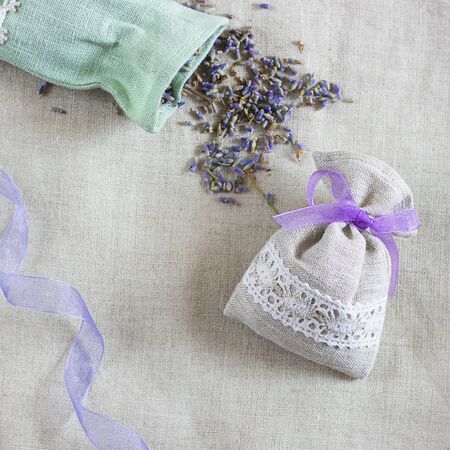 Small linen sack filled with dried lavender decorated with lacework and violet ribbon coqueand and one sachet is opened. Top view on thenatural flaxen background. Square with copy spaceの写真素材