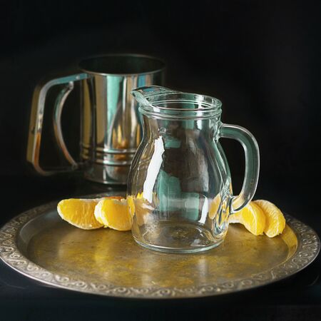 Glass decaner and metal mug and several bright segments of orange on vintage tray on dark background. Square. Still life composition imitation of visual art painting.の写真素材
