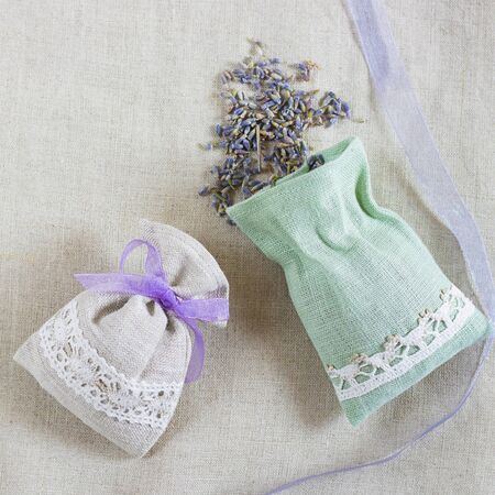 Small linen bag filled with dried lavender decorated with lacework and violet ribbon coque, two sachets, one is opened. Square with copy space. Top view on thenatural flaxen background.の写真素材