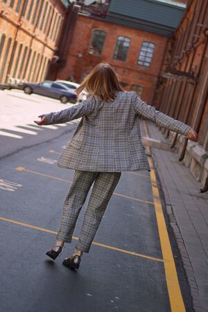 Unrecognizable blond lady wearing plaiting trouser suit going and jumping among the street with red brick buildings in positive mood.の写真素材