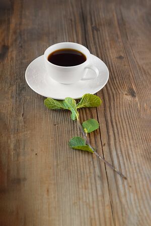 elegant white cup of hot drink in porcelain tea-set on brown rustic wooden table and fresh green mint sprig put together like flower. Creative food concept. Open composition. Vertical with copy space.の写真素材