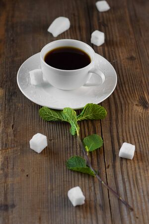 white porcelain tea-set with hot drink in on brown rustic wooden table with fresh green mint sprig put together like flower surrounded by pieces of sugar . Creative food concept. Vertical, copy spaceの写真素材
