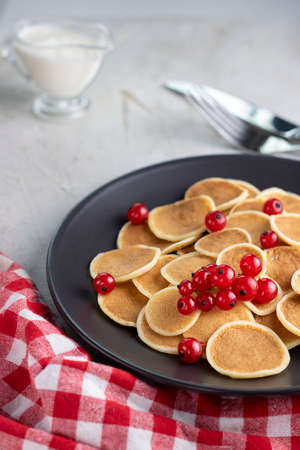 Tiny pancake cereal on black plate served with red currant berries, sauce, utensils on grey table with gingham tablecloth. Trendy breakfast, round baked crepe batter drops. Homemade food. Vertical.の写真素材