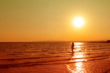 Dark silhouette of young woman seen from far away, going alone by the sea on the background of sunset. Orange tone. Horizontal with copy space. Holiday concept.の写真素材