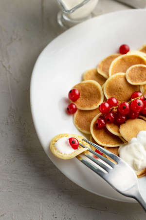 Tiny pancake cereal on white plate served with red currant berries and ingredients on grey cement table. Trendy breakfast, small round baked crepe batter drops. Homemade food. Verticalの写真素材