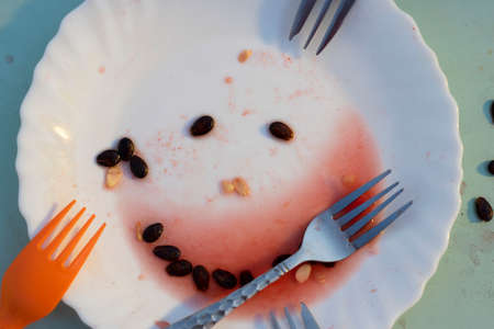 Kid background. image of all that remains of whatermelon on white plate. Dish with black seeds put as smile with forks around. Summer party for children. Horizontal food concept.の写真素材