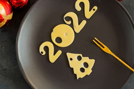 Figures 2022 and furtree made of cheese with holes, on a black plate with small fork and Christmas tree decoration, symbol of proper nutrition, menu. Horizontal. Winter and New year conceptの写真素材