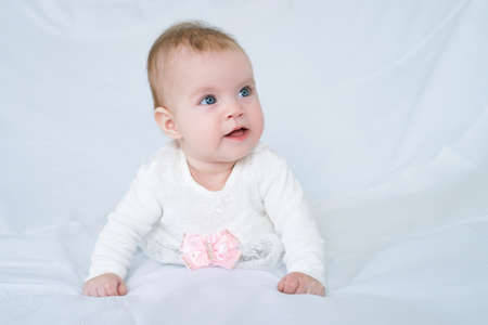 Cute baby with blue eyes in white dress with pink bow lying on her stomach looking at camera, white background,の写真素材