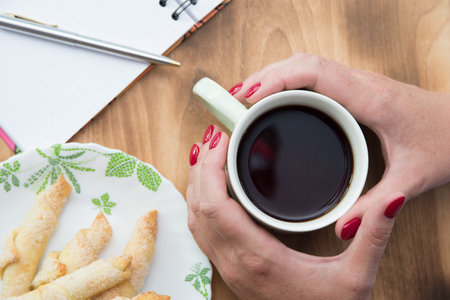 Woman hand holding a cup wooden background. Cookie, ballpoint pen and empty notepad with copy space...