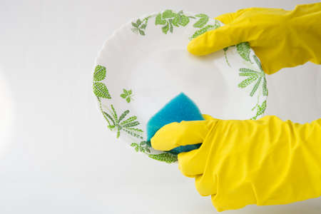 Close up of female hands in yellow protective rubber gloves washing white plate with blue cleaning sponge against white backgroundの写真素材