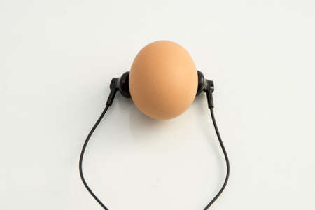 small black headphones lie next to the chicken egg as if the egg is listening to music, white background orange eggの写真素材
