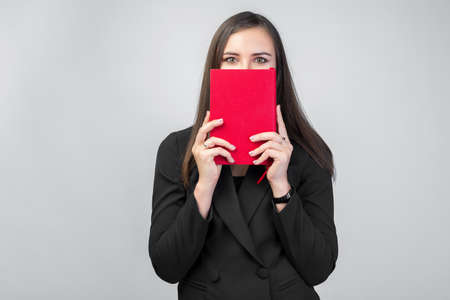 a young brunette girl in a business suit holds a red notebook in front of her face, white background, dark straight hair, only beautiful eyes are visibleの写真素材