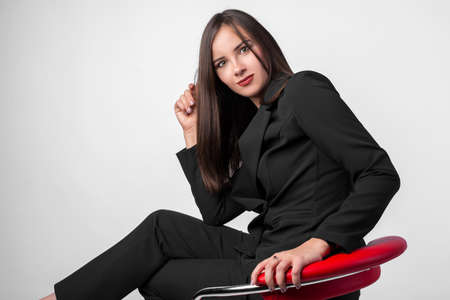 Young beautiful girl in business suit black jacket, pants, shoes sitting on red chair, white background, business lady, young cute brunetteの写真素材