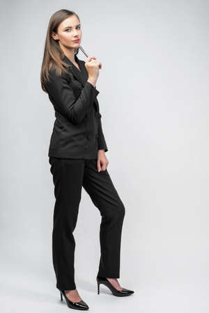 young girl in a black business suit holding a ballpoint pen in her hands, white background, pants, jacket, black high-heeled shoes, business ladyの写真素材