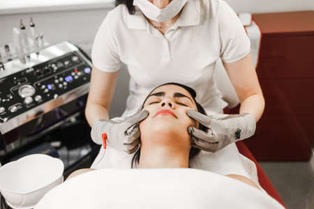 Microcurrent therapy. Cosmelotologist makes stimulating facial massage with electric gloves. Lymphatic drainage massageの写真素材