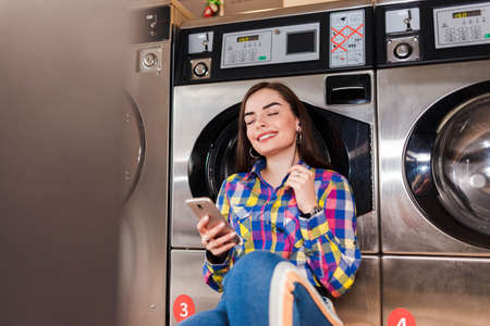 Pretty girl with smartphone in hand listens to music leaning on a washing machine in public laundryの写真素材