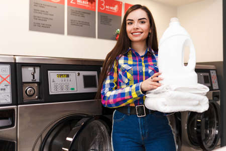 Satisfied girl in the launderette holds a stack of white towels and laundry detergentの写真素材