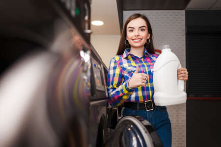 Young woman recommends a detergent holding a bottle in her hand and showing thumb up. Woman with washing detegrent bottle in public laundryの写真素材