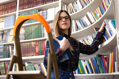 Cute student in glasses takes a book from the library bookshelf. Young lady is looking for the right literature among many booksの写真素材