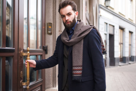 Business elegant man opens the door to his office. Stylish man in a coat with a beard enters the...