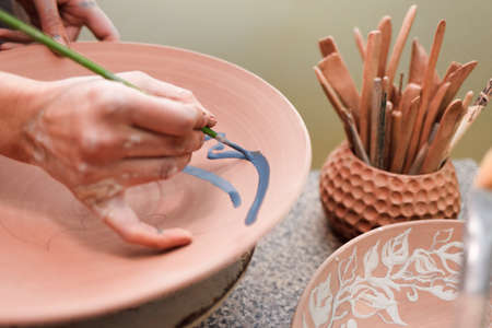 Potter woman paints a ceramic plate closeup. Girl draws with a brush on earthenware. Process of creating clay products.の写真素材
