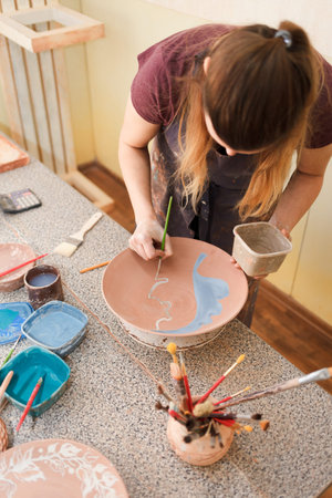 Potter woman paints a ceramic plate. Girl draws with a brush on earthenware. Process of creating...