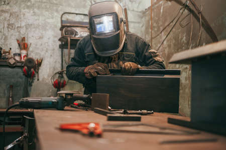 Worker in a welder mask works in a workshop for welding iron. Man makes iron products. Guy works with a welding machine using a mask to protect his eyes from dangerous raysの写真素材
