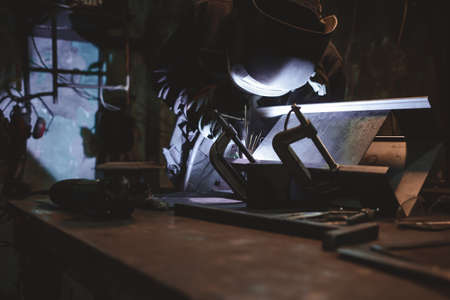 Worker in a welder mask works in a workshop for welding iron. Man makes iron products. Guy works with a welding machine using a mask to protect his eyes from dangerous raysの写真素材