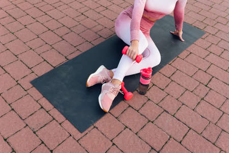 Girl with perfect figure in beautiful pink sportswear sits on yoga mat with dumbbell in handsの写真素材