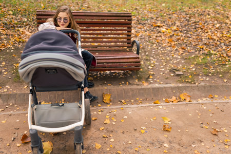 Mom with baby carriage sits on bench in park and looks on the baby who sleeps in stroller