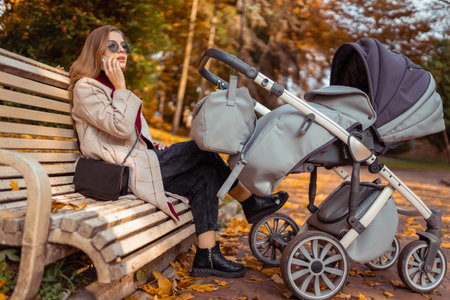 Stylish mom with stroller talking on the phone while sitting on bench in the park while the child is...