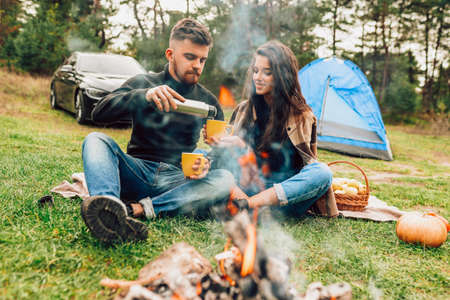 Man pours his girlfriend tea from vacuum flask while sitting in the nature near campfireの写真素材