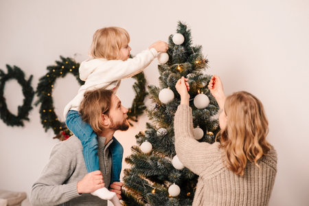Happy family with child decorate christmas tree. Little girl sitting on daddy's shoulders hangs ball on the Christmas treeの写真素材