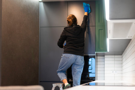 Woman stands on stepladder and wipes kitchen cabinets with rag. Kitchen cleaning process