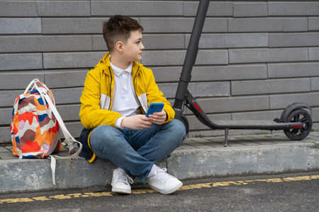 Teenager with mobile phone sitting next to his electric scooter and backpack. Modern young boyの写真素材