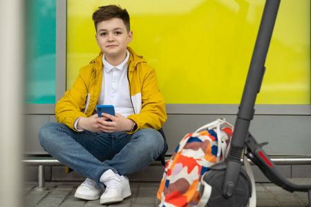 Teenager with mobile phone sitting next to his electric scooter and backpack. Modern young boyの写真素材