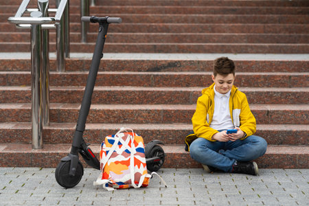 Modern teenager sitting on the stairs near his electric scooter and texting on smartphoneの写真素材