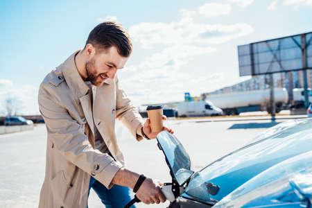 Stylish man with coffe cup in hand inserts plug into the electric car charging socketの写真素材