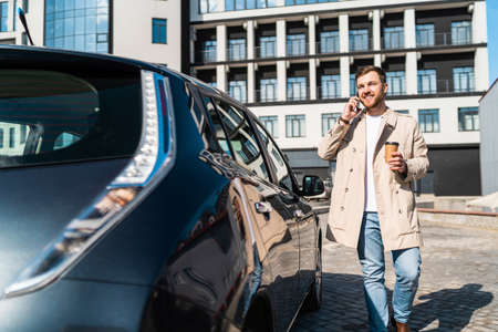 Handsome man with cup of coffee in his hand talking on the phone while standing near his carの写真素材