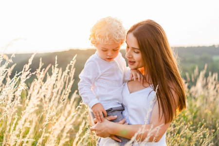Beautyful woman with a little curly-haired boy in her arms stands in the middle of a field at sunset