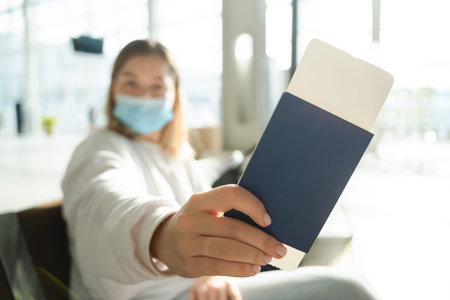 Young woman in a mask sits in an airport lounge during a pandemic and shows her passport and ticket...