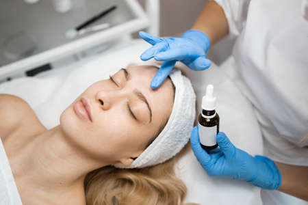 Beautician vitamin serum oil on face applying skin of beautiful girl. Young blonde woman during spa treatments in a beauty salonの写真素材