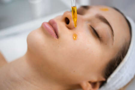 Close up of beautiful woman receiving nourishing oil treatment in beauty salon. Beautician holding pipette and applying serum on face of woman in spa salonの写真素材