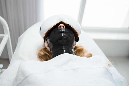 Blonde woman with a black alginate mask on her face and eye massage glasses lies on a couch of a beauty salon. Spa procedures and relaxationの写真素材