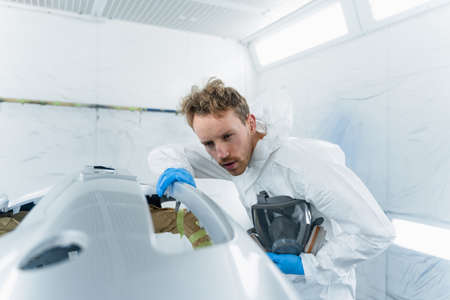 Young car painter examines the surface of the car bumper in detail for the quality of the paintwork performed. Auto service employee works in the spray boothの写真素材