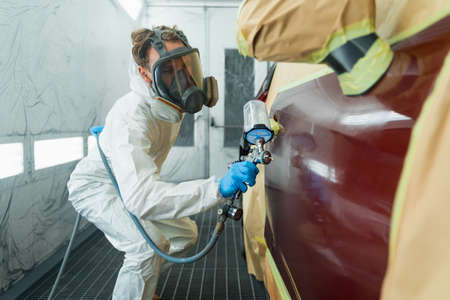 Auto painter in a protective suit and mask sprays a degreaser or other cleaner on a car door using a spray gun. Preparation of vehicle body elements for local repairの写真素材