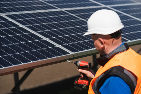 Male electrical engineer in a hard hat with electric wrench maintains solar panels at power plant