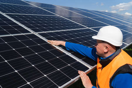 Solar power plant worker in hard hat checks the surface of the photovoltaic panel with his hand for damageの写真素材